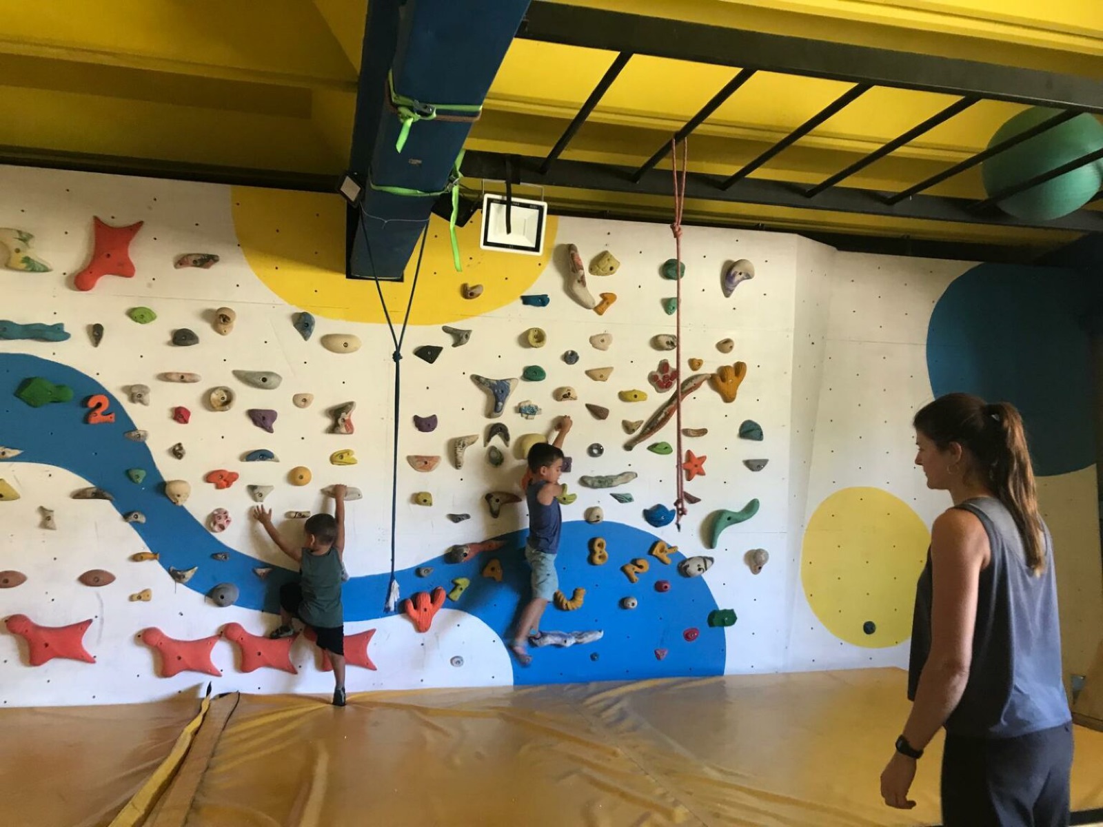 Due bambini arrampicano su una parete boulder blu, seguiti da un'istruttrice